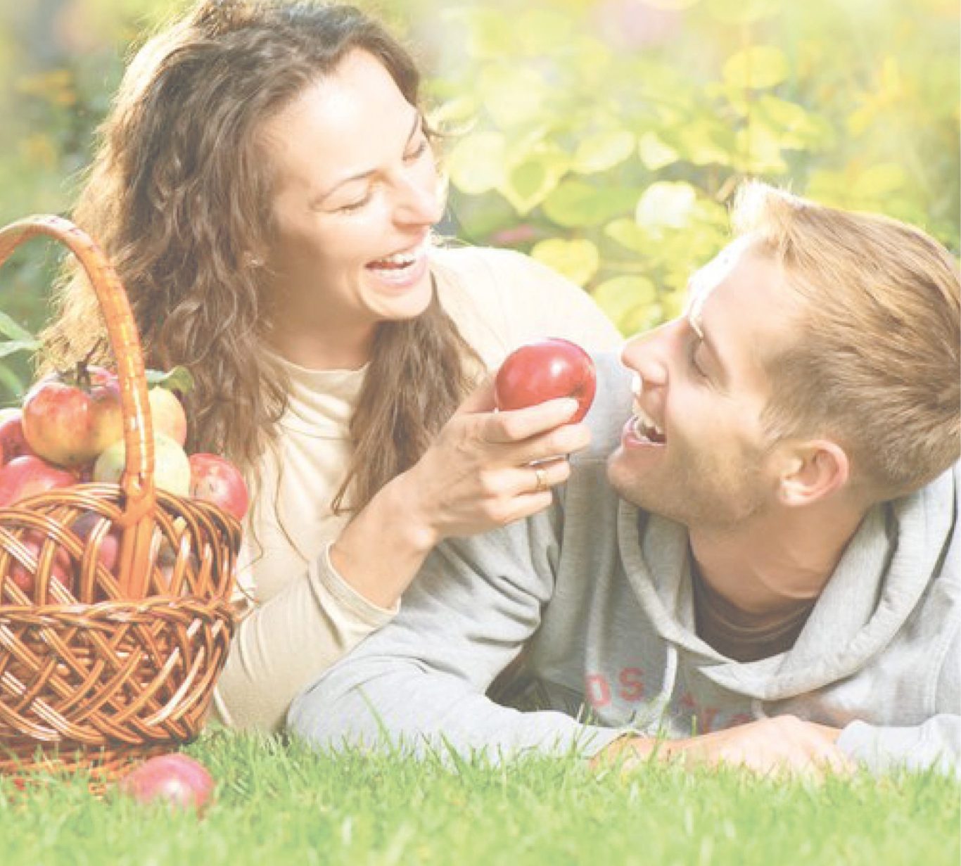 Frau hält einen Apfel, lächelt mit Mann in einem blühenden Garten. Korb mit Äpfeln daneben.